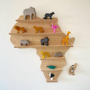 Może przedstawiać: Drewniana półka w kształcie Afryki, prezentująca różne drewniane figurki zwierząt. Zwierzęta obejmują słonia, lwa, żyrafę i nosorożca. Półka wykonana jest z jasnego drewna i zamontowana na białej ścianie.