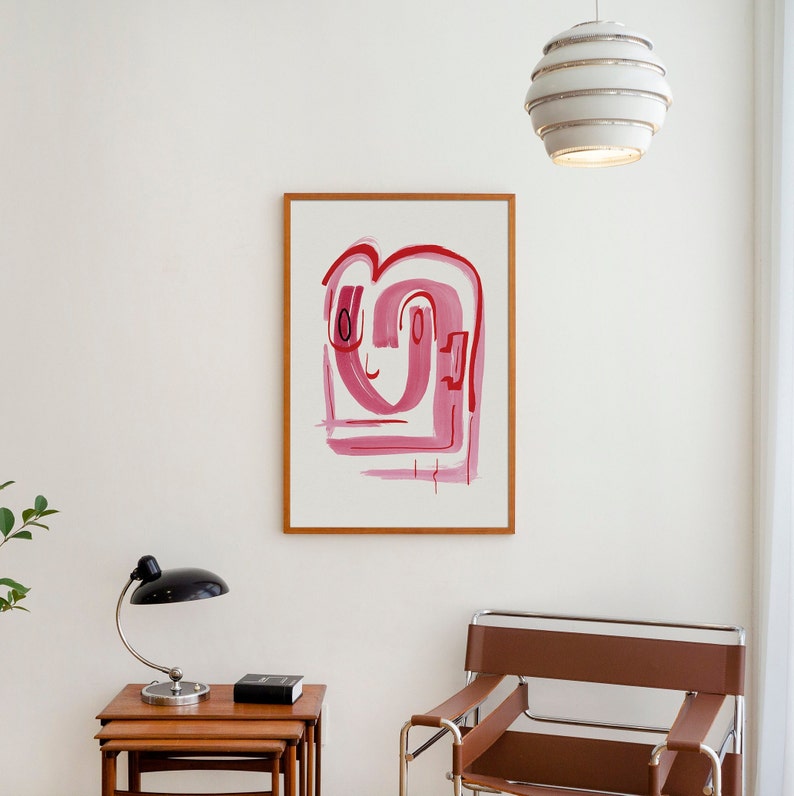 May include: A framed abstract painting with pink and red brushstrokes on a white background. The painting is hanging on a white wall in a room with a chair and a lamp.
