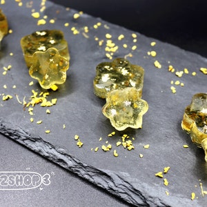 May include: Close-up of several star-shaped, golden-yellow candies with gold flakes, arranged on a dark gray slate board. The candies are translucent, and small yellow particles are scattered around them. The image has a high-angle perspective.