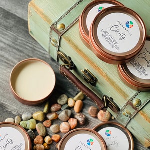 May include: Several copper-coloured tins of "Clarity" all-natural deodorant balm, each containing 71g. The tins are placed near a vintage green trunk and pebbles, with the product label visible.