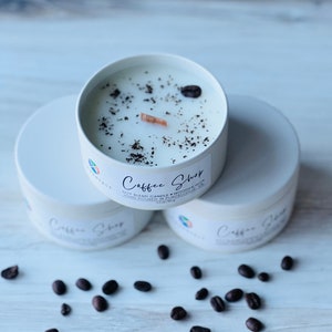 May include: Three white soy blend candles with a wooden wick. The candles are labeled "Coffee Shop" and have coffee beans and a wooden wick on top. The candles are sitting on a white surface with coffee beans scattered around them.