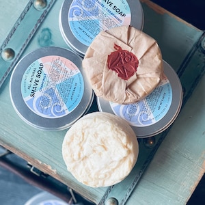 May include: Four round containers of shave soap, two in silver tins with labels that read "All-Natural Shave Soap" and "Chamomile", one in a silver tin with a label that reads "Lavender + Cedarwood Shave Soap", and one wrapped in brown paper with a red wax seal.
