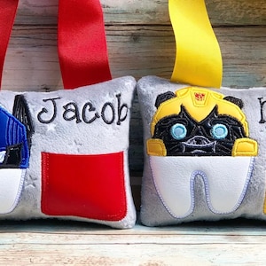 May include: Two gray tooth pillows with embroidered Transformers characters. The pillow on the left has a blue and black Optimus Prime character and the text "Jacob". The pillow on the right has a yellow and black Bumblebee character and the text "Mason".