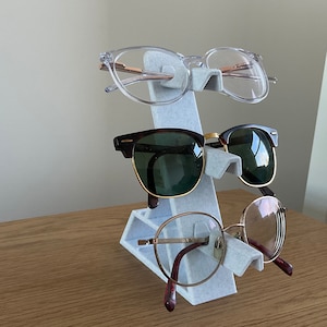 May include: A white plastic stand with three slots for holding spectacles. The stand is holding three pairs of glasses: one clear pair, one brown pair with green lenses, and one brown pair with clear lenses.