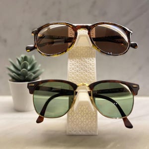 May include: Two pairs of sunglasses on a white stand. The top pair has brown tortoise shell frames and brown lenses. The bottom pair has dark brown frames and green lenses.