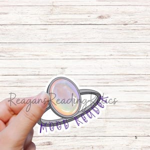 May include: A sticker featuring a ring design with a large, oval-shaped, iridescent gem. The ring is silver-colored, and the sticker has the text "Mood Reads" and "Reagans Reading Rics" in purple and light gray.