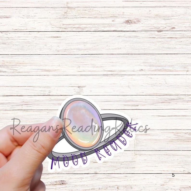 May include: A sticker with a ring design, featuring a large, oval-shaped, iridescent gem. The ring is silver-coloured, and the sticker has the text "Mood Reads" and "Reagans Reading Rics" in purple and light grey.