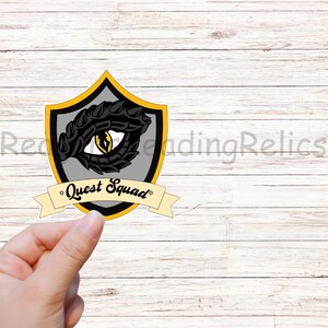 May include: A yellow and gray shield sticker with a black dragon eye and the text "Quest Squad" on a white wood background.