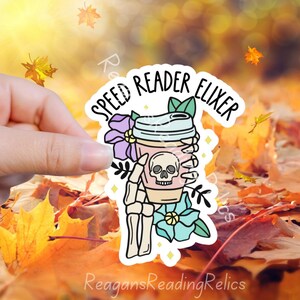 May include: A sticker with a skeleton hand holding a pink and white coffee cup with the text "Speed Reader Elixir" in a white circle with a floral design.