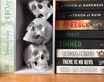 May include: Three silver skull figurines stacked inside a white box on a wooden shelf. The box is in front of a bookshelf with several books with titles like "A Touch of Darkness", "Flock", "Priest", "Sinner", "Brutal Prince", "There is No Devil", and "There Are No Saints".