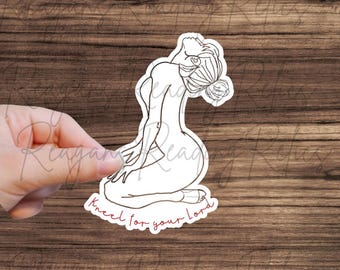 May include: A white sticker featuring a line drawing of a person kneeling with their head tilted back. The text "Kneel for your lord" is written in red script below the figure. The sticker is held against a wooden background.