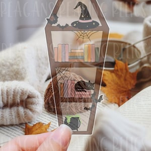 May include: A transparent coffin-shaped bookmark featuring a printed illustration of two shelves. The top shelf displays a witch hat and books, while the bottom shelf shows books, a black cat, and a spider web. The text Transparent Bookmark is visible.
