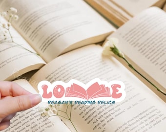 May include: A pink and white sticker with the words "LOVE REAGAN'S READING RELICS" and a pink open book illustration. The sticker is on a book with white flowers.