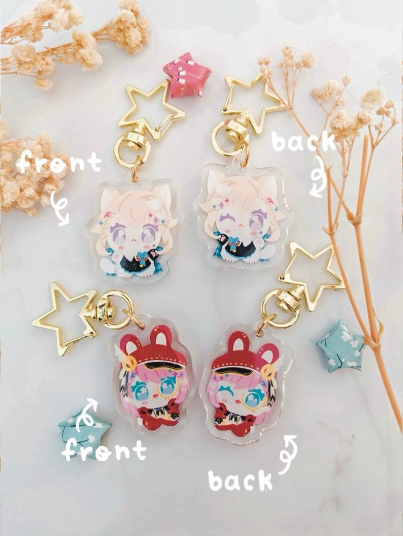 K&ouml;nnte beinhalten: Transparente Acryl-Schl&uuml;sselanh&auml;nger mit Anime-Charakteren und goldenen sternf&ouml;rmigen Verschl&uuml;ssen. Ein Design hat einen Charakter mit hellem Haar und einem schwarz-blauen Outfit. Das andere Design hat einen Charakter mit rosa Haaren und einem roten Outfit.