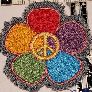 May include: A colorful flower-shaped patch with a peace sign in the center. The flower has five petals in red, yellow, green, blue, and purple. The patch is made of denim and has a frayed edge.