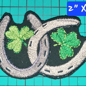 May include: Embroidered patch featuring two intertwined horseshoes and two green shamrocks. The patch is approximately 2 inches by 2 inches. The horseshoes are silver, and the shamrocks are green on a black background.