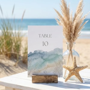 May include: A beach-themed table setting with a white table, a sign that reads "TABLE 10", a starfish, and a vase with dried pampas grass. The sign has a watercolor design in shades of blue and green. The background is a beach with the ocean.