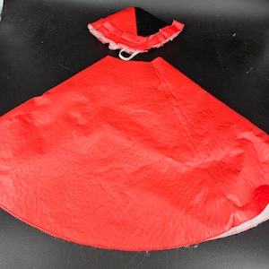 Op de afbeelding: Een rood en zwart kostuum met een cape en een hoed. De cape is gemaakt van rode stof en heeft een witte rand. De hoed is zwart met een rode rand en een witte ruche.