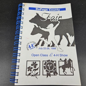 Op de afbeelding: Een wit boekje met een blauwe en witte titel "DuPage County Fair" met een silhouet van een meisje dat over een koe springt. Het boekje is de 44e jaarlijkse beurs en vindt plaats van 22 tot en met 26 juli 1998. De tekst "Open Class & 4-H Show" staat onder de afbeelding. Het boekje heeft een blauwe spiraalbinding.