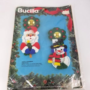 Puede incluir: Un kit de lona de plástico para hacer dos cubiertas para pomos de puerta. Una cubierta presenta a un Papá Noel sosteniendo un árbol de Navidad y la otra presenta a un muñeco de nieve. El kit incluye hilo acrílico, una aguja e instrucciones. El paquete está etiquetado como "Santa & Snowman" y "Bucilla".