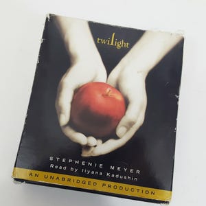 Puede incluir: Una caja rectangular de audiolibro para Twilight de Stephenie Meyer, narrado por Ilyana Kadushin. La portada muestra un par de manos sosteniendo una manzana roja brillante sobre un fondo negro, con el título en amarillo y una etiqueta de producción íntegra.
