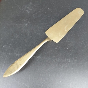 May include: A gold-colored cake server with a long, slender handle and a wide, flat blade. The server is made of metal and has a polished finish. The handle tapers to a point, and the blade is rectangular with rounded corners. The server is designed for serving cakes and pastries.