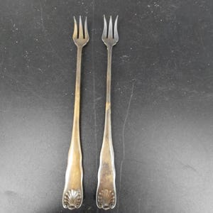 Pode incluir: Dois garfos de cocktail em metal prateado com cabo longo, apresentando três dentes finos e um motivo decorativo de concha em relevo na base de cada cabo. Os garfos têm um acabamento liso e ligeiramente manchado.