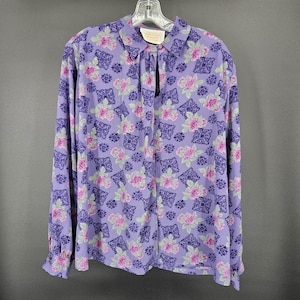 May include: A long-sleeved lavender blouse with a floral pattern of pink roses and dark purple geometric shapes. The blouse has a collared neckline and button closure. The sleeves have a slight ruffle at the cuffs.