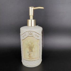May include: A decorative soap dispenser with a cream-colored, textured body and a gold-colored pump. The front features a framed image of a potted plant. The dispenser has a tag attached.
