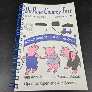 Op de afbeelding: Een spiraalgebonden boek met een blauwe spiraalrug. De omslag toont de tekst "Du Page County Fair" en "JULY 23-27, 2003". Drie cartoonvarkens dansen onder de tekst "SOMETHING TO SQUEAL ABOUT!". Het boek is getiteld "49th Annual Premium Book".