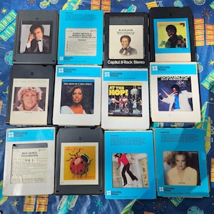 May include: A collection of vintage 8-track tapes featuring various artists. The tapes have black and white cases with blue and white labels. Album covers include portraits and graphics. The collection is from Columbia House.