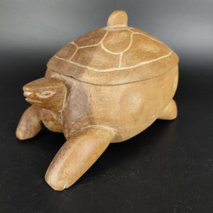 Könnte beinhalten: Eine geschnitzte Holzbox in Schildkrötenform mit Deckel. Die Schildkröte ist hellbraun mit einem detaillierten Schalenmuster und einem kleinen Knopf oben. Die Box hat vier Beine und einen Schildkrötenkopf.