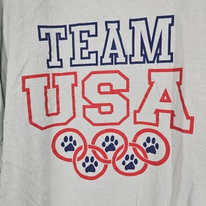 May include: A light gray sweatshirt with the words "TEAM USA" in blue and red block letters. Below the text are five interlocking red circles, each containing a blue paw print.