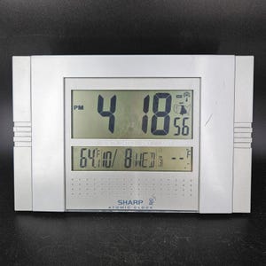Puede incluir: Un reloj atómico Sharp plateado que muestra la hora 4:18 PM y 56 segundos. La pantalla inferior muestra la fecha, el día y la temperatura a 17.7 grados Celsius. El reloj tiene forma rectangular.