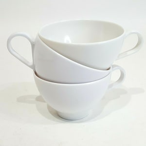 May include: Two stacked white ceramic cups with smooth, rounded bowls and curved handles. The top cup rests at an angle within the lower cup, creating a layered appearance. Both pieces feature a clean, solid white finish with no patterns.