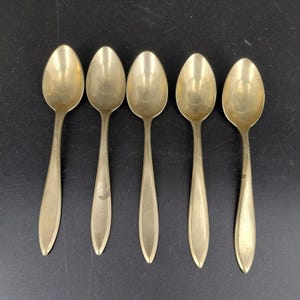 May include: Five vintage gold-colored spoons arranged in a row. Each spoon has a rounded bowl and a slender, elongated handle. The spoons are made of a shiny metal, possibly brass or gold-plated, and are set against a dark background.