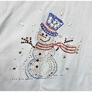 May include: A light blue top featuring a snowman design made of rhinestones. The snowman wears a red and white striped scarf and a blue hat with white stars. Surrounding the snowman are scattered star-shaped rhinestones.