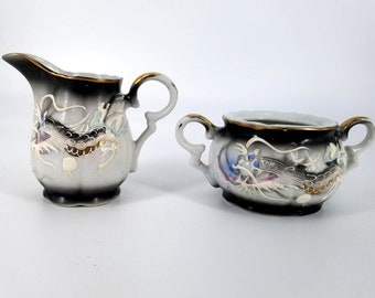 Op de afbeelding: Een set van een witte porseleinen roomkan en suikerpot met een zwart en gouden drakenontwerp. De draak is geschilderd in tinten blauw, groen en goud.