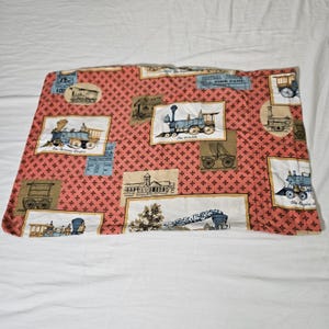 May include: A coral-colored fabric featuring a repeating pattern of small brown crosses. The fabric is decorated with framed illustrations of vintage trains in various styles and settings. The illustrations are in shades of blue, brown, and white.