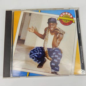 Puede incluir: Una portada de CD con un hombre que lleva una camisa con estampado azul y blanco y un sombrero azul, sosteniendo una serpiente. La portada tiene un fondo amarillo con el texto "Cobra Bad Boy Talk" en un círculo rojo.