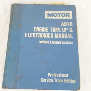Puede incluir: Un manual azul MOTOR Auto Engine Tune Up & Electronics, Professional Service Trade Edition. La portada presenta texto blanco, incluyendo "Includes Emission Servicing". El libro muestra signos de desgaste.