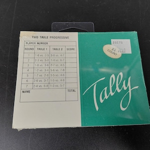 May include: A Two Table Progressive tally card set in a teal and white package. The front features a printed grid for player numbers, rounds, table assignments, and scores. The word Tally is printed in white script, and a sticker reads 24 COUNT.