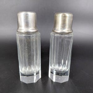 May include: Two clear glass salt and pepper shakers with silver-colored metal lids. The shakers have a rectangular shape with vertical grooves and sit on a faceted, hexagonal base. The lids are smooth and cylindrical.