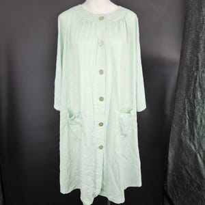 May include: A light green, button-down robe with long sleeves and two front pockets. The robe has a round neckline and is made of a soft, flowing fabric. The buttons are a matching light green color.