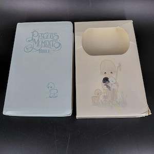 May include: A light blue "Precious Moments Bible" with matching box. The book cover features the title in script and a lamb illustration. The box has a cutout and a pastoral scene with a shepherd and lambs.