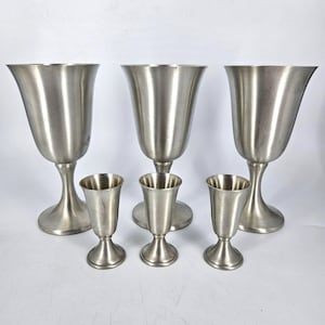 May include: A collection of six brushed metal goblets and shot glasses. The three larger goblets have a flared top and a rounded base. The three smaller shot glasses have a straight, cylindrical shape. All pieces have a silver finish.