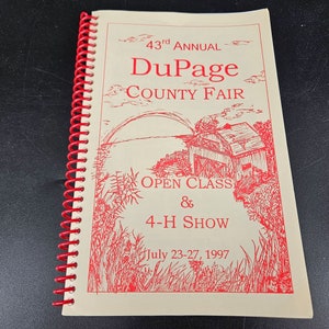 Op de afbeelding: Een rood en wit programma voor de 43e jaarlijkse DuPage County Fair. Het programma bevat een tekening van een rode schuur met een regenboog in de lucht. De tekst "Open Class & 4-H Show" is onder de schuur gedrukt. De data van de beurs zijn onder de tekst gedrukt: "July 23-27, 1997".