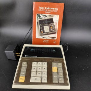 May include: A vintage Texas Instruments TI-5100 electronic calculator with an orange manual. The calculator has a white and brown body with a display showing numbers. The manual reads "Texas Instruments electronic calculator TI-5100".