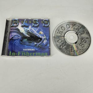 Puede incluir: Una caja de CD y un disco con el título "Hooked on Bass" con "The In-Fisherman". La caja tiene un fondo azul con un gráfico de pez. El disco es plateado con el título impreso.