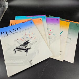 May include: A set of six piano lesson books, titled "Piano Adventures", with colorful covers featuring musical notes and a piano with wings. The books are numbered 24, 2B, 3B, 1, and 5.
